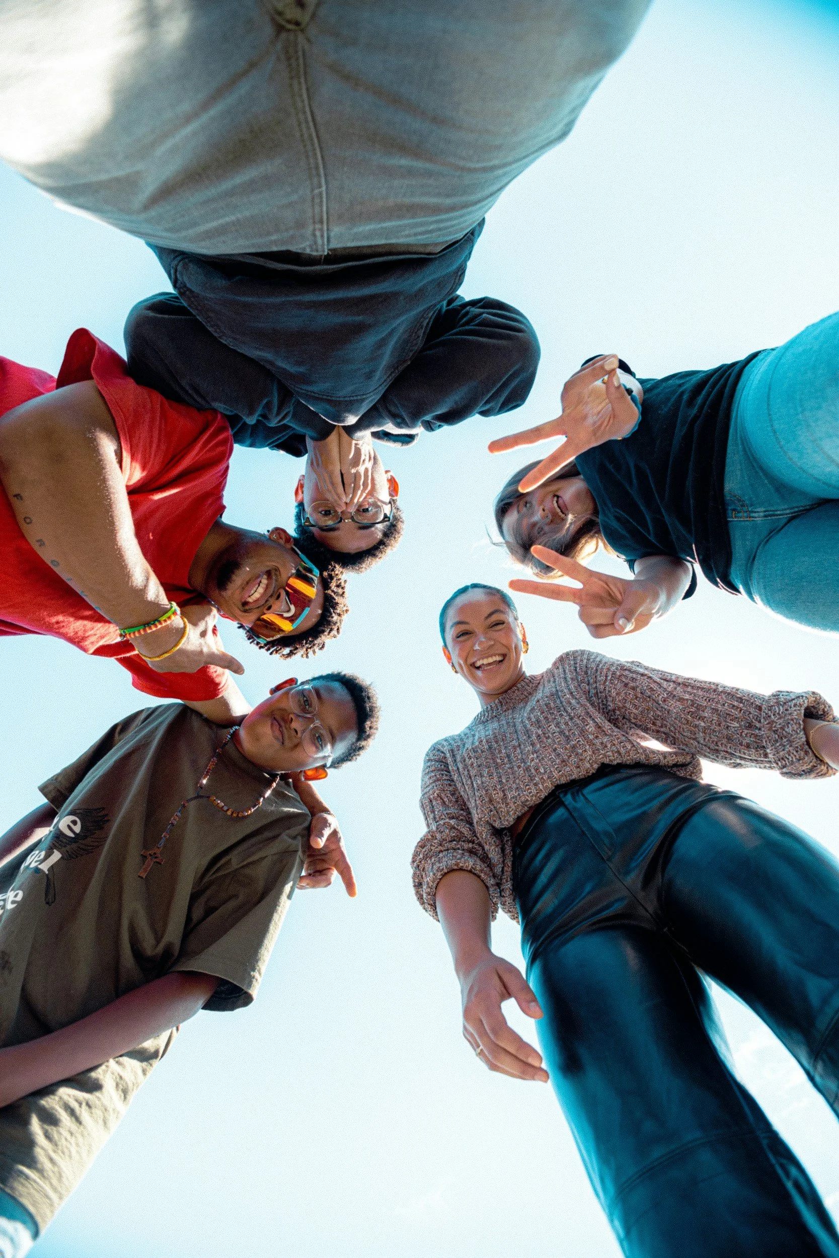 Group of diverse young people standing in a circle, looking down at the camera, smiling and making peace signs, outdoors under a clear blue sky.