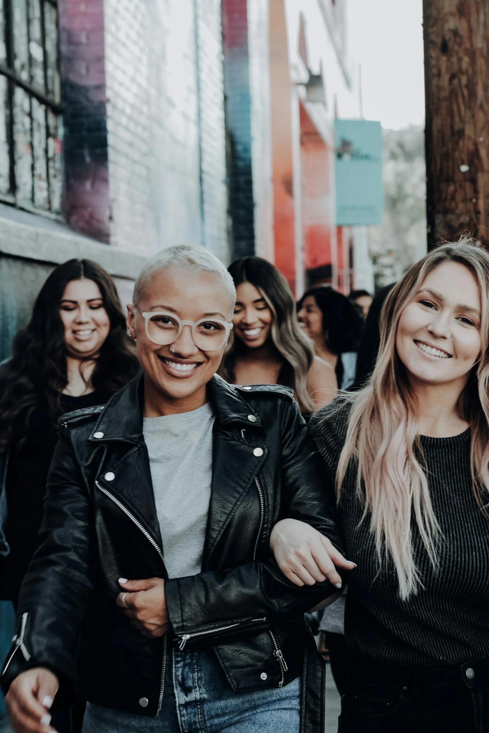 Group of smiling women walking outdoors on a city street, holding each other’s arms, with colorful buildings in the background.