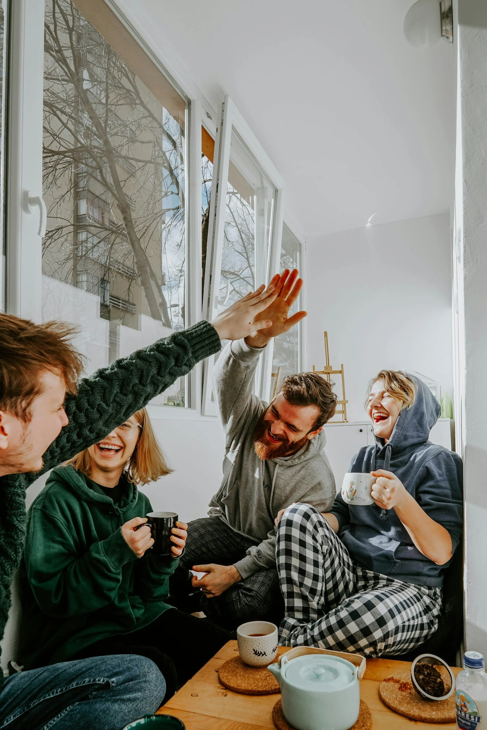 Group of five young adults sitting on the floor, enjoying coffee and laughing together in a bright room with large windows.