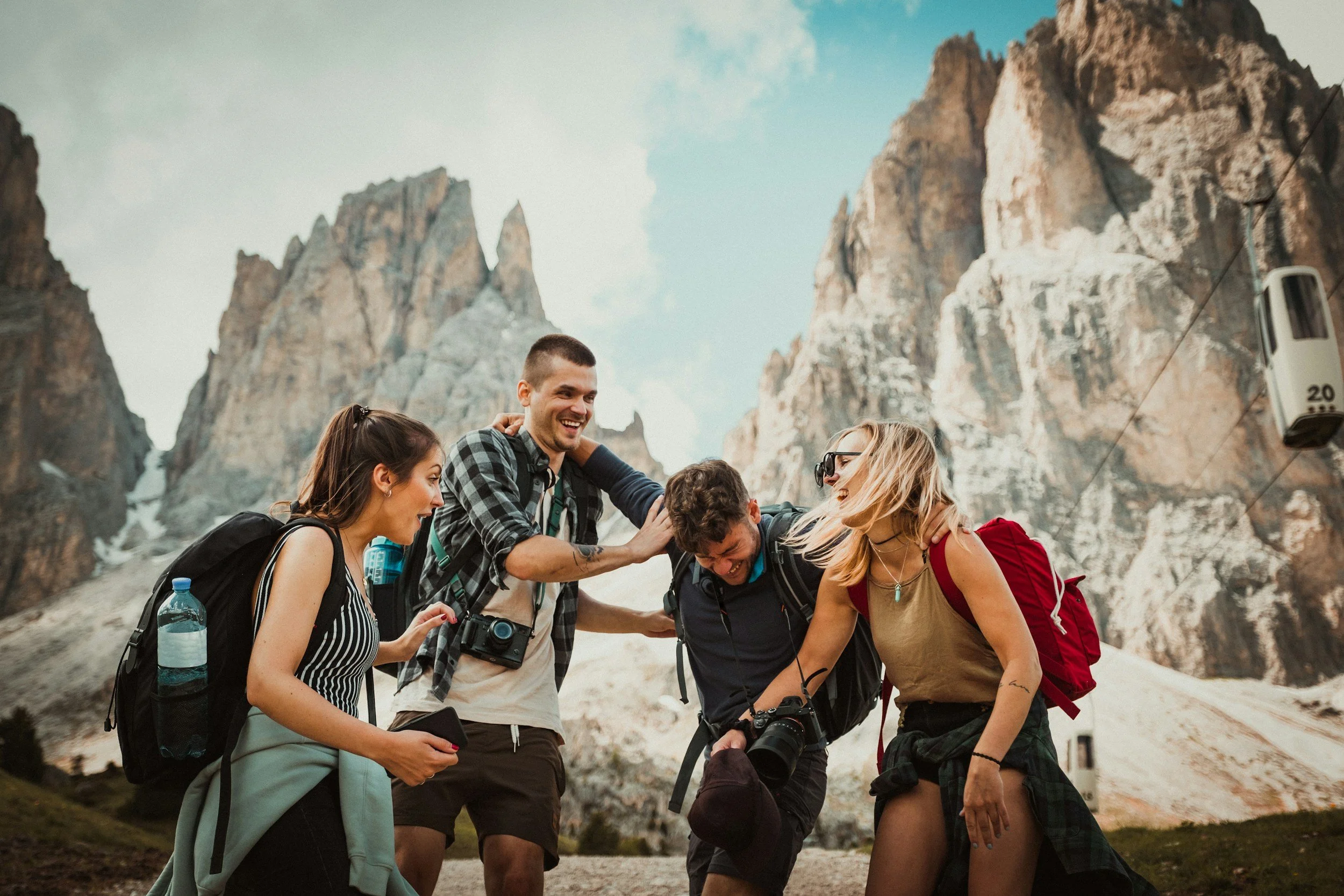 Four friends, two women and two men, laughing and joking together while hiking in a mountainous area with rocky peaks in the background. They are dressed in casual outdoor clothing, carrying backpacks, and holding cameras.